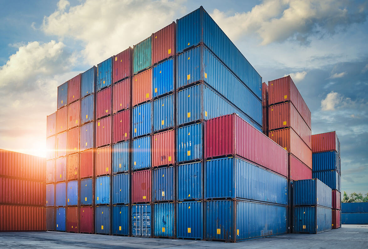 Stacks of shipping containers on a dock with the sun rising behind them