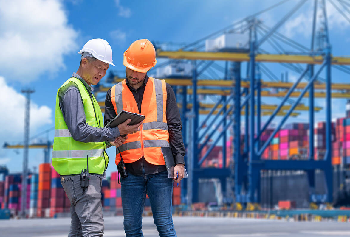 Two dock workers dressed in safety gear with a tablet and container cranes in the background